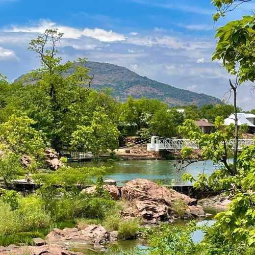 A scenic view of a river surrounded by lush greenery and rocky formations, with mountains in the background under a blue sky.