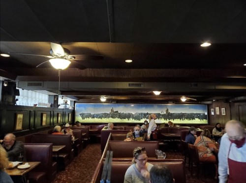 Interior of a restaurant with booths, diners, and a mural of cowboys on horseback in the background.