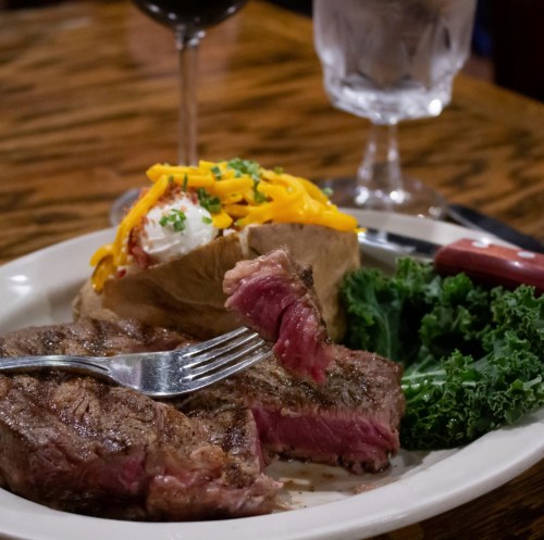 A plate with a juicy steak, a loaded baked potato, and kale, accompanied by a glass of water and red wine.