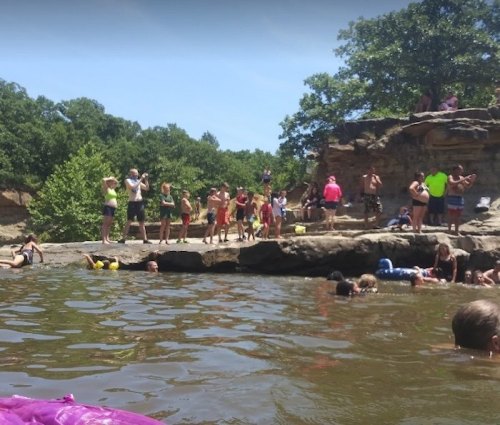 A lively scene at a swimming area with people enjoying the water and sun on a rocky shore surrounded by trees.