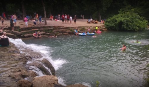 A lively river scene with people swimming, lounging, and enjoying the water on a sunny day. Green trees in the background.