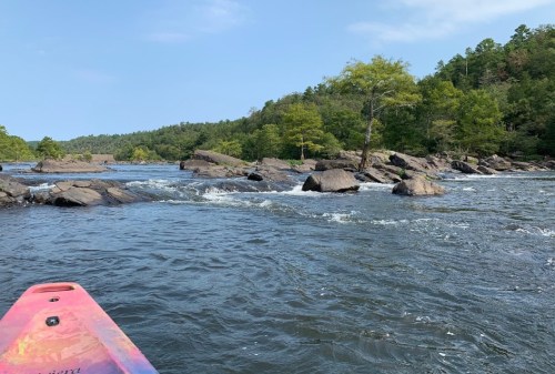 A view of a river with rocky outcrops and lush green trees along the banks, seen from a kayak.