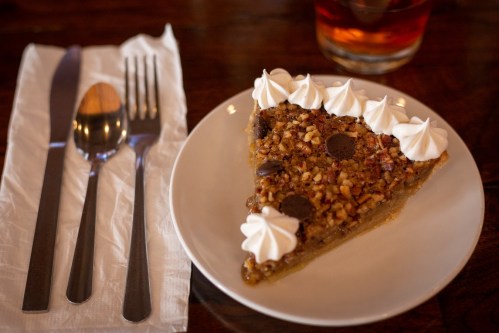 A slice of pecan pie topped with whipped cream, served on a plate with utensils and a drink in the background.