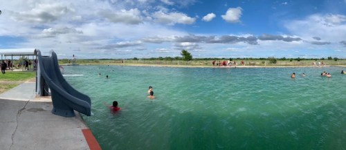 A panoramic view of a swimming area with a slide, people swimming, and a cloudy sky in the background.
