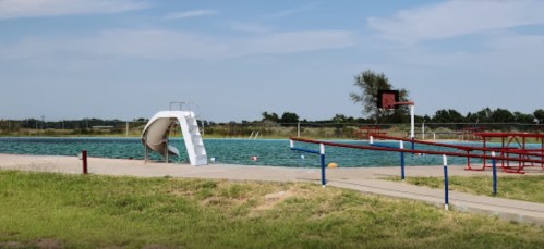 A swimming pool with a slide, surrounded by grass and a basketball hoop in the background under a clear blue sky.