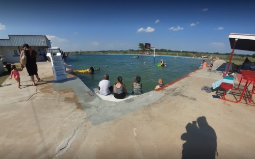 A group of people relax by a pool, with some swimming and others sitting on the edge, under a clear blue sky.