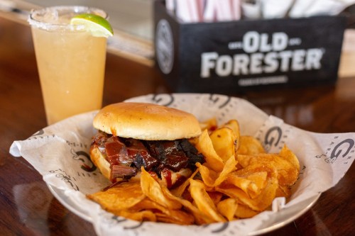 A BBQ sandwich with brisket and sauce, served with crispy chips and a cocktail with a lime wedge.