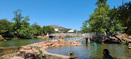 A sunny park scene with people swimming and a bridge over a calm lake, surrounded by trees and rocky shores.