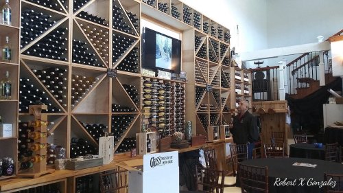 A wine cellar with wooden racks filled with bottles, a staircase in the background, and a man standing near a table.