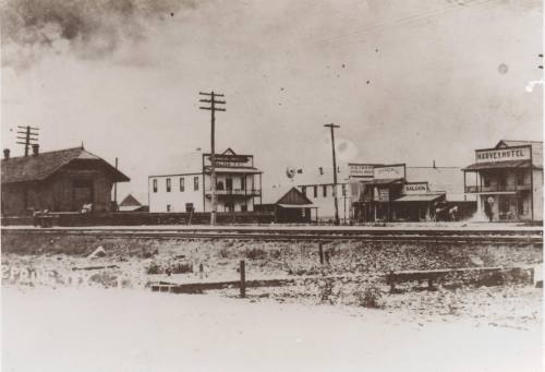 Historic black-and-white photo of a small town with buildings, a hotel, and a saloon near railroad tracks.