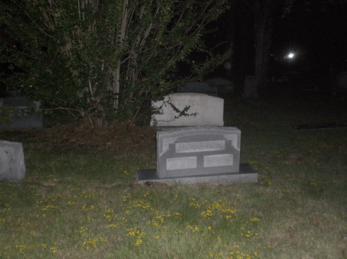 A dark cemetery scene featuring a gravestone with the name "Houston" surrounded by grass and flowers.