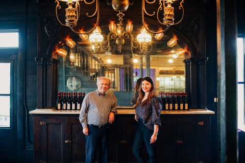 A man and woman stand in front of a vintage bar with wine bottles, illuminated by ornate hanging lights.