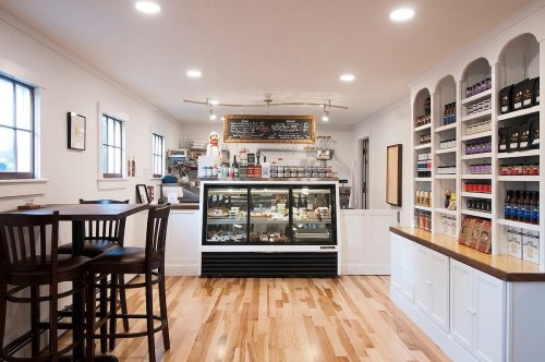A cozy café interior featuring a display case, shelves of products, and a small seating area with wooden chairs.