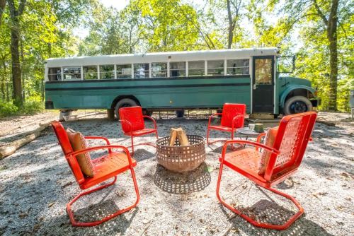 A green converted bus parked in a wooded area, surrounded by four red chairs and a fire pit on gravel.