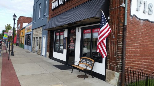 A storefront with an American flag, featuring signs for oysters and shrimp, next to colorful buildings on a sidewalk.