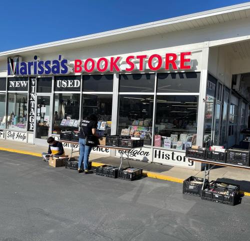 A bookstore exterior with large signage; two people organize books outside on tables under clear blue skies.
