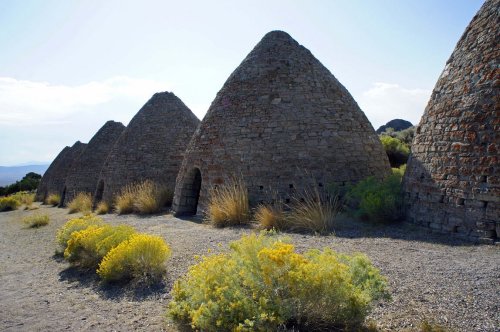 Row of stone beehive-shaped kilns surrounded by dry grass and shrubs under a clear blue sky.