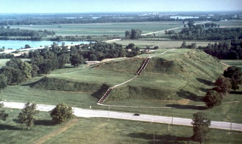Aerial view of a large, grassy mound with steps, surrounded by trees and fields, near a road and water.