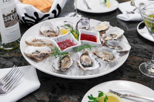 A plate of fresh oysters on ice, served with lemon wedges and two sauces, alongside drinks and bread in a restaurant setting.