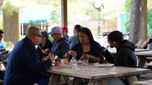 A group of people dining at picnic tables under a shelter, enjoying food and drinks in a casual outdoor setting.