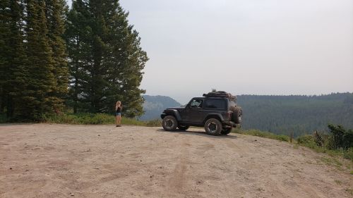 A person stands near a parked off-road vehicle on a dirt road, surrounded by tall trees and a misty mountain view.