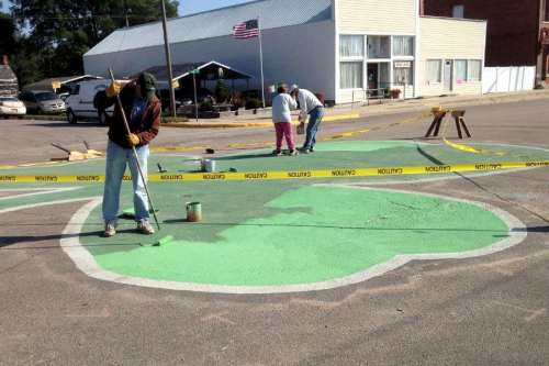 People painting a large green shamrock on a street, with caution tape and a building in the background.