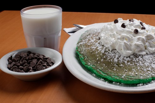 A green pancake topped with whipped cream and chocolate chips, served with a glass of milk and a bowl of chocolate chips.