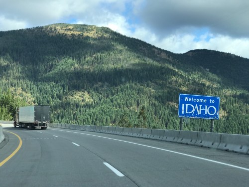 A truck drives past a "Welcome to Idaho" sign on a highway surrounded by green mountains and cloudy skies.