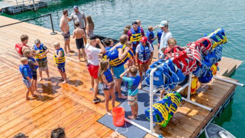 A group of children and adults on a dock, preparing to put on life jackets by the water.