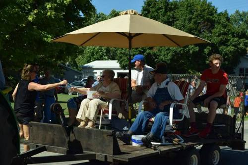 A group of people sitting under an umbrella on a trailer, enjoying a parade, with one person handing out refreshments.