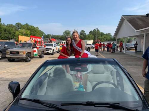 Two young women in red dresses sit on a convertible car, smiling at a parade with festive decorations in the background.