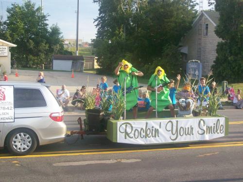 A parade float featuring performers in corn costumes, promoting "Rockin' Your Smile," with spectators in the background.