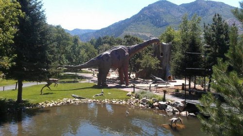 A dinosaur sculpture in a park, surrounded by trees and a pond, with mountains in the background.