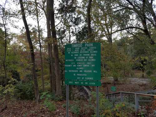 A green sign at Timrod Park outlines rules for vehicle access and park usage, surrounded by trees and autumn foliage.