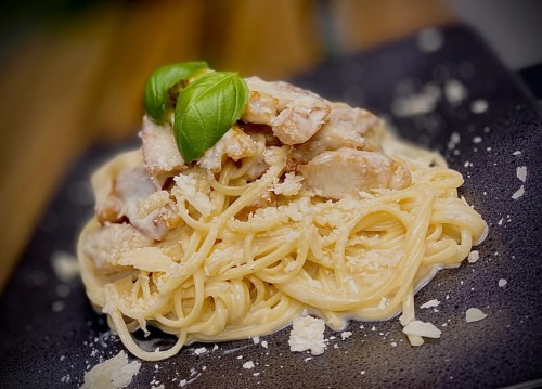 A plate of creamy pasta topped with chicken, basil, and grated cheese, served on a dark plate.