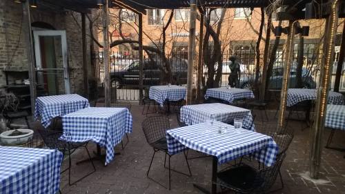 Outdoor dining area with checkered blue and white tablecloths, surrounded by bare trees and a brick pathway.