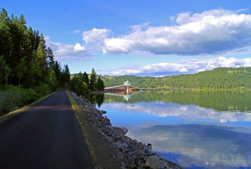 A serene lake surrounded by trees, with a path alongside and a bridge reflecting in the calm water under a blue sky.