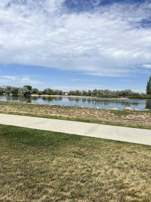 A serene landscape featuring a calm lake, grassy shore, and a cloudy blue sky in the background.