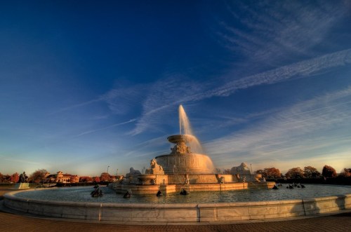 A large, ornate fountain with a tall water jet, surrounded by a serene park under a clear blue sky.