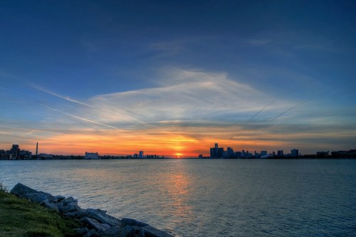 Sunset over a city skyline, reflecting on calm waters, with colorful clouds in the sky and rocky shoreline in the foreground.
