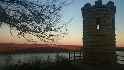 A stone tower overlooks a river at sunset, surrounded by trees with autumn leaves. Soft colors fill the sky.
