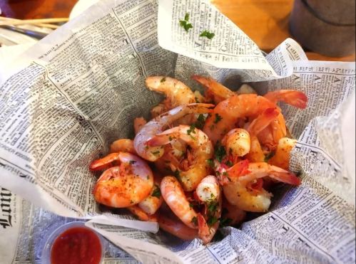 A serving of shrimp seasoned and garnished, presented in a paper cone with a side of dipping sauce.