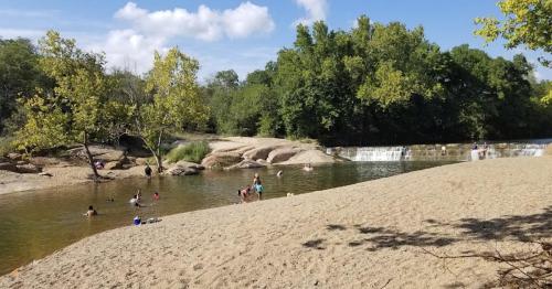 A sandy beach by a river with people swimming and playing, surrounded by trees and a small waterfall in the background.