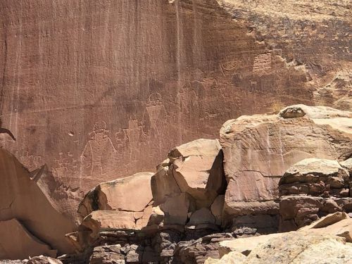 Petroglyphs carved into a rocky cliff, depicting human figures and symbols, surrounded by large boulders.