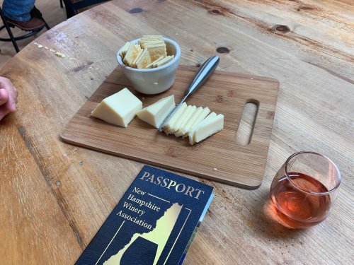 A wooden board with sliced cheese, a bowl of crackers, and a glass of rosé wine next to a passport for a winery association.