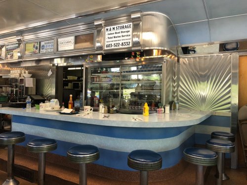 A retro diner counter with blue and silver decor, bar stools, and a menu board above. Condiments are on the counter.