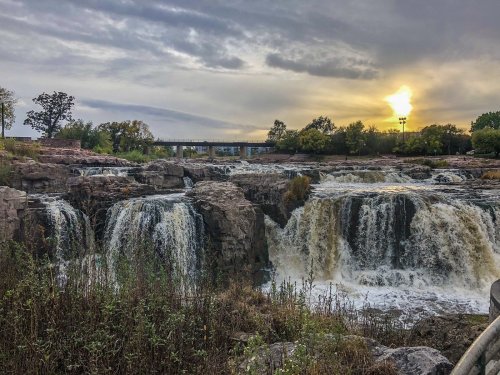 A scenic view of waterfalls at sunset, with rocky formations and lush greenery in the foreground.