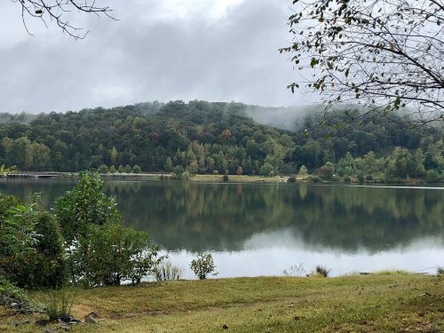 A serene lake surrounded by lush green hills, reflecting the cloudy sky and trees in calm waters.