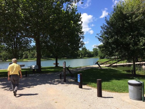 A man walks along a path by a serene lake surrounded by trees and a clear blue sky.