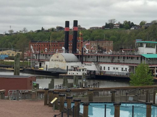 A historic steamboat, the William M. Black, docked by a riverside with green hills and buildings in the background.